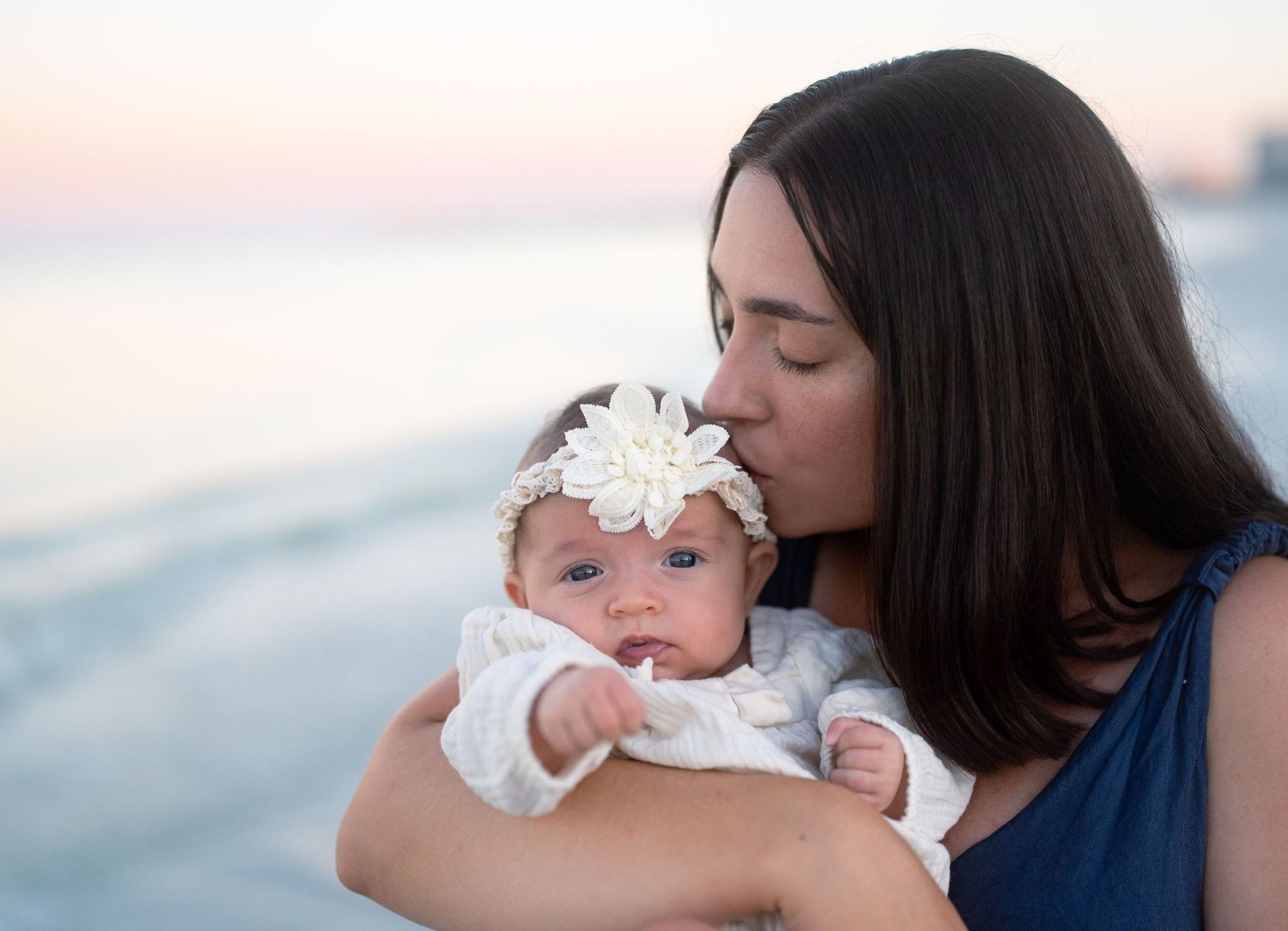 Tender moment captured at sunset as mother kisses baby wearing white floral headband.
