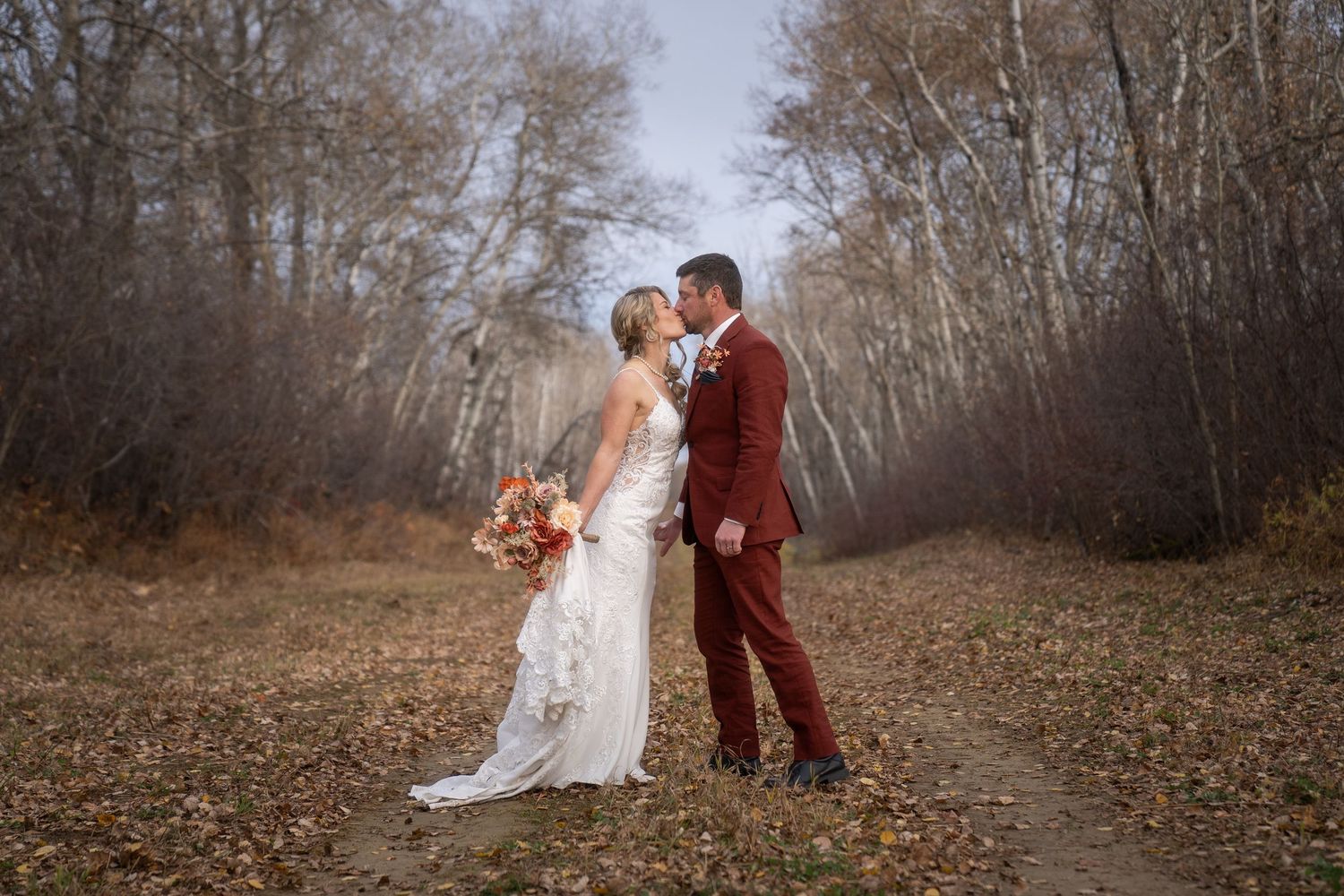 A couple in wedding attire holding hands while walking down a tree-lined path during autumn.