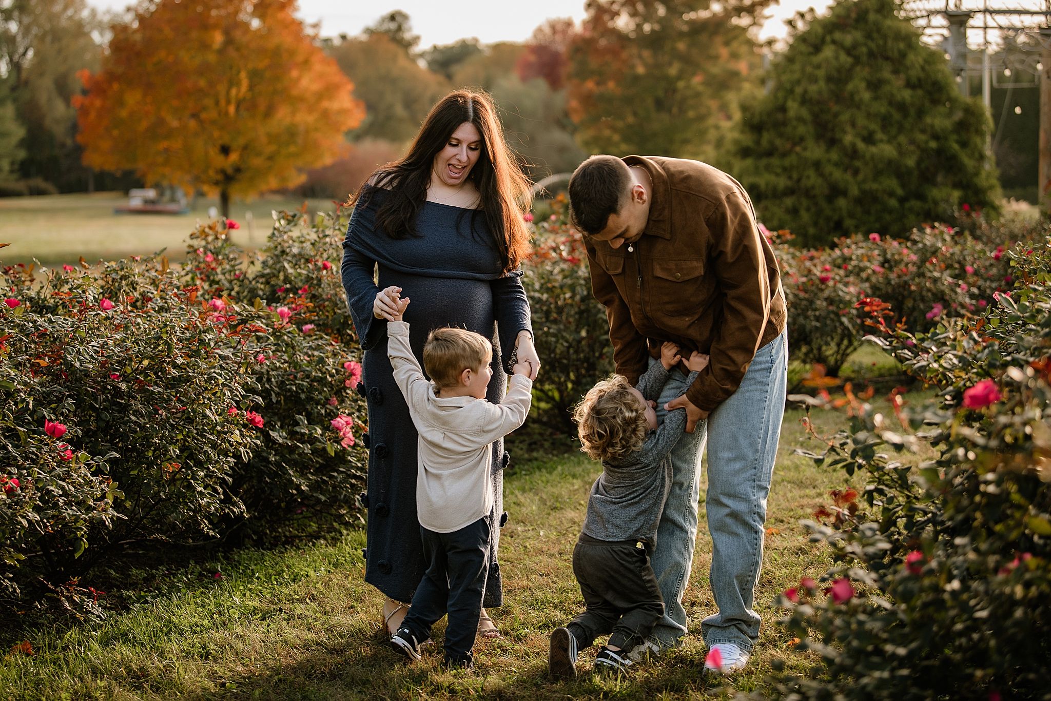A family plays together in a garden of blooming roses during autumn with colorful fall foliage in the background.