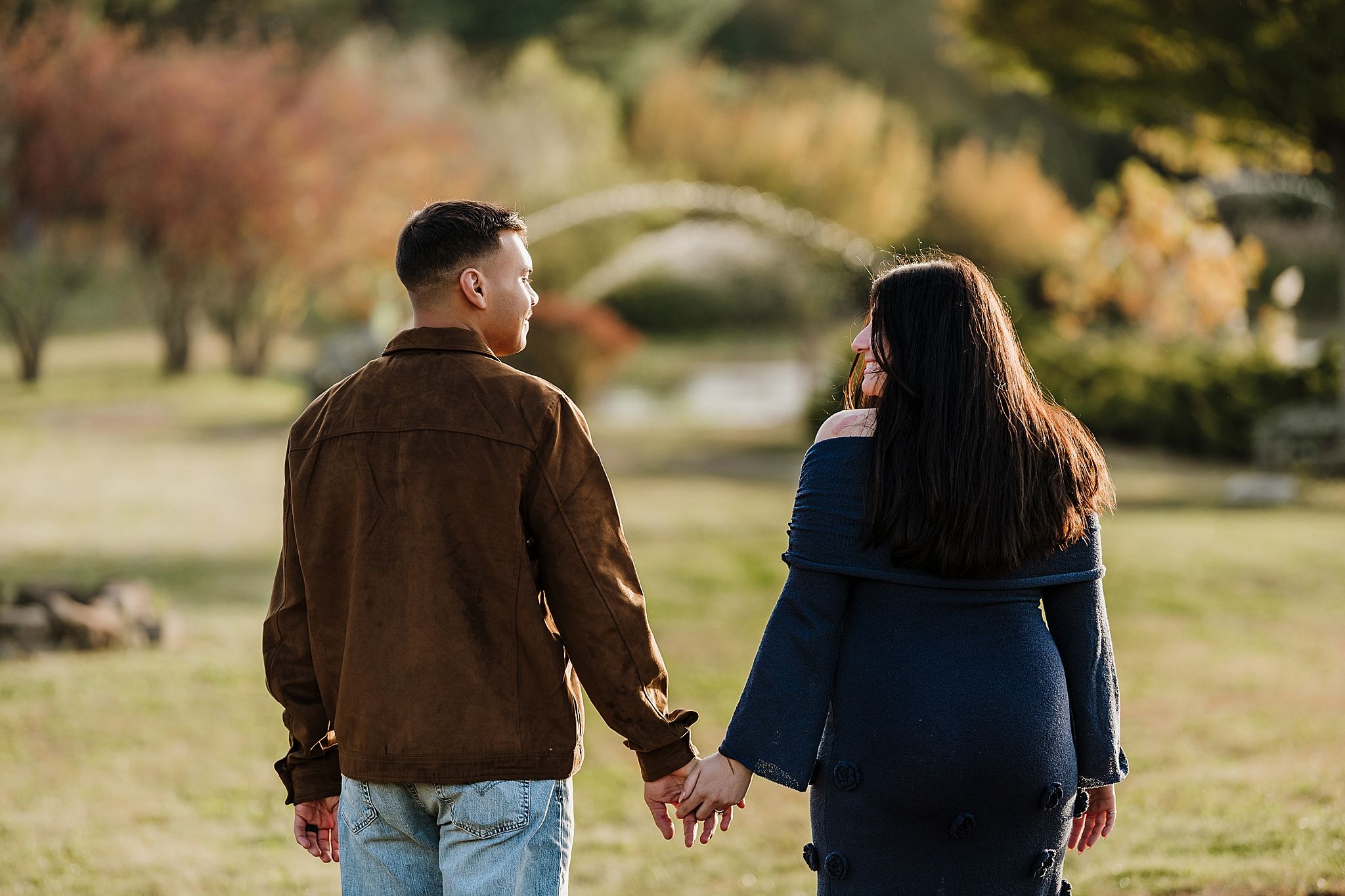 A couple walks hand in hand through a sunny garden setting with autumn foliage in the background.
