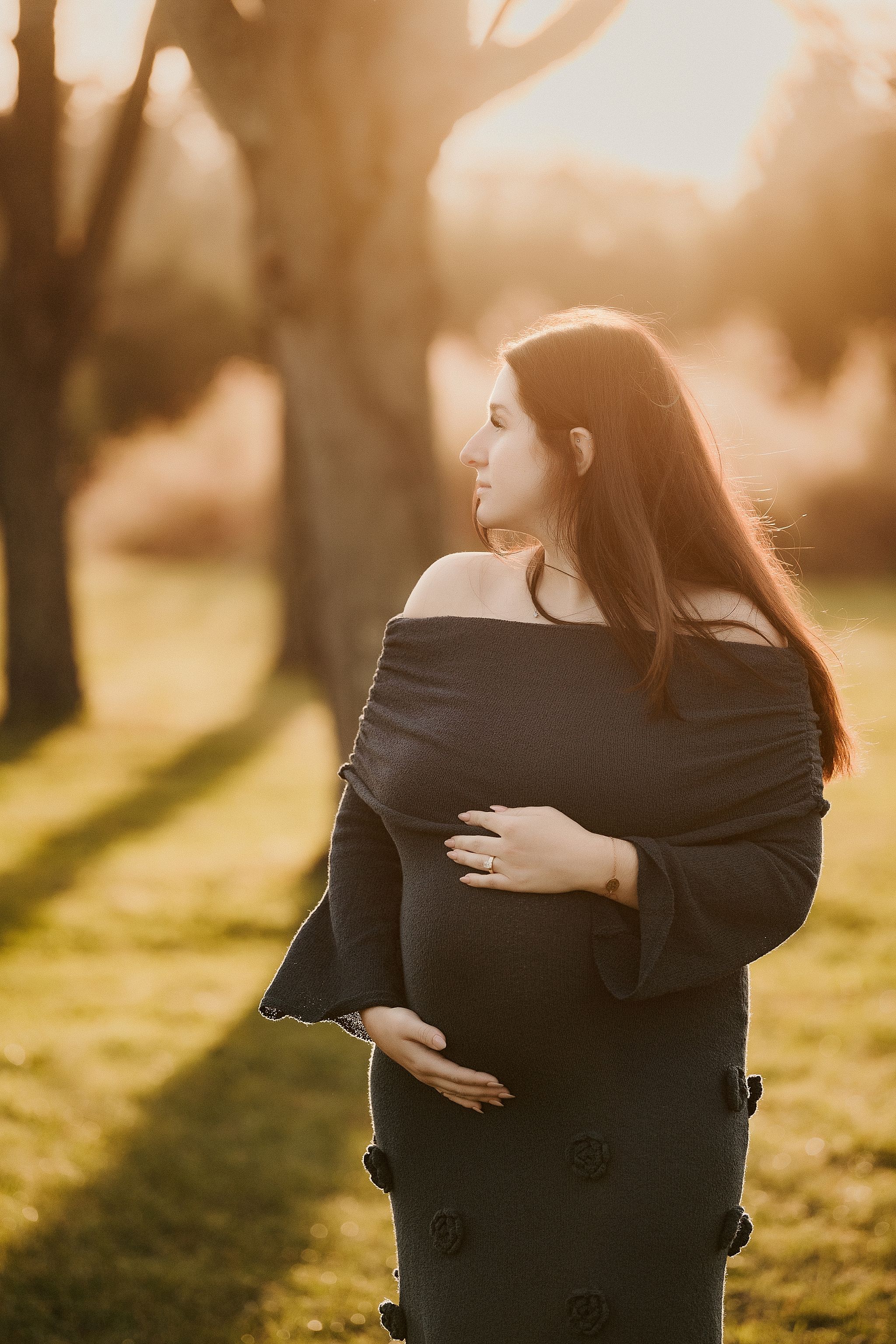 Silhouetted pregnant woman in black dress stands in golden sunlight during outdoor maternity photoshoot.