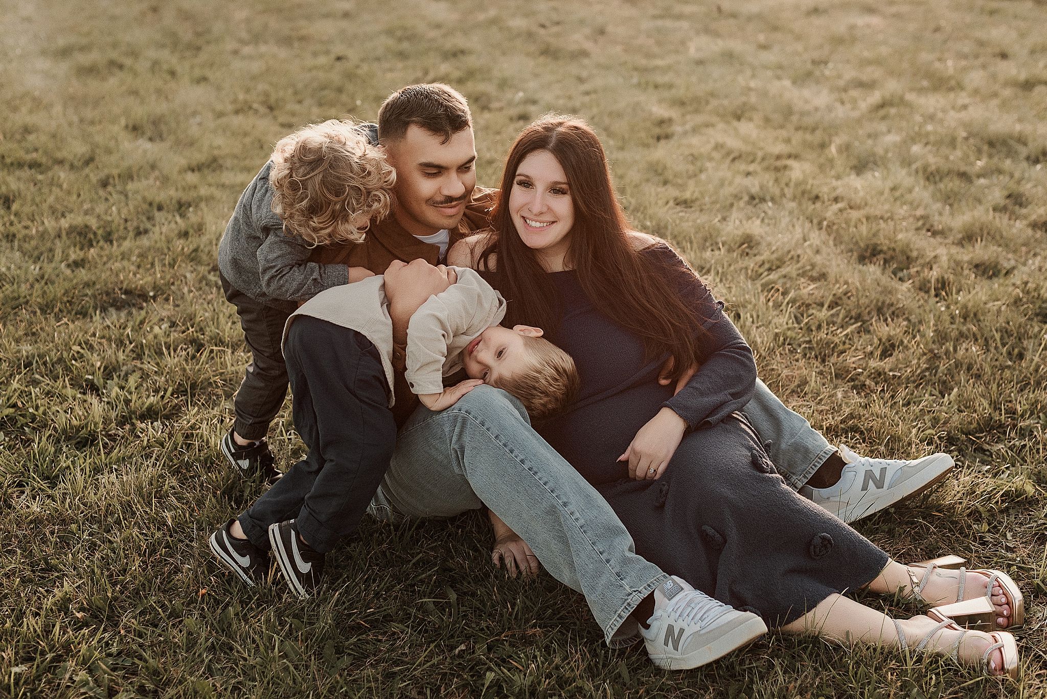 A couple sits together on grassy hillside during golden hour sharing intimate moments in casual attire.