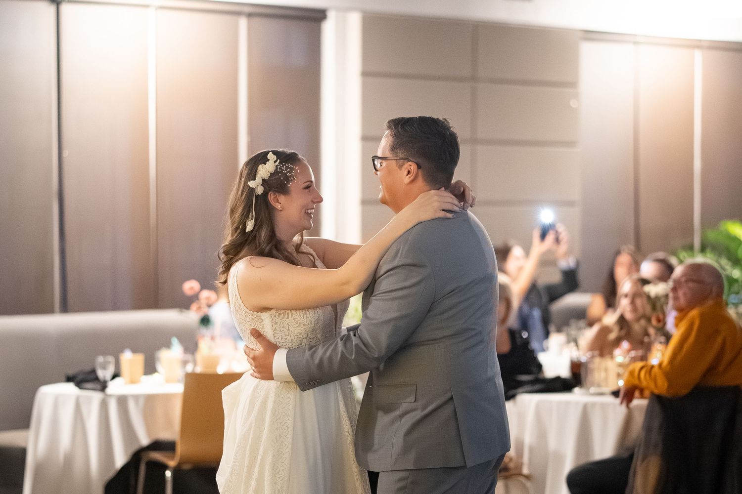A couple shares their first dance at their wedding reception while guests look on from tables in the background.