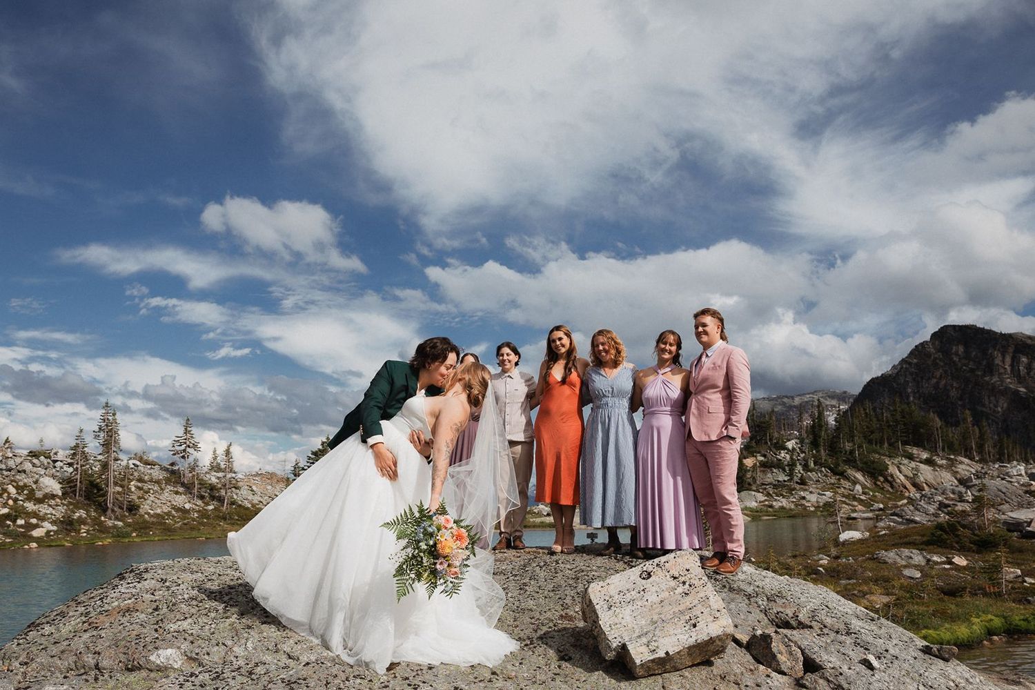 Bride and wedding party pose on rocky alpine lake shore under dramatic cloudy skies.