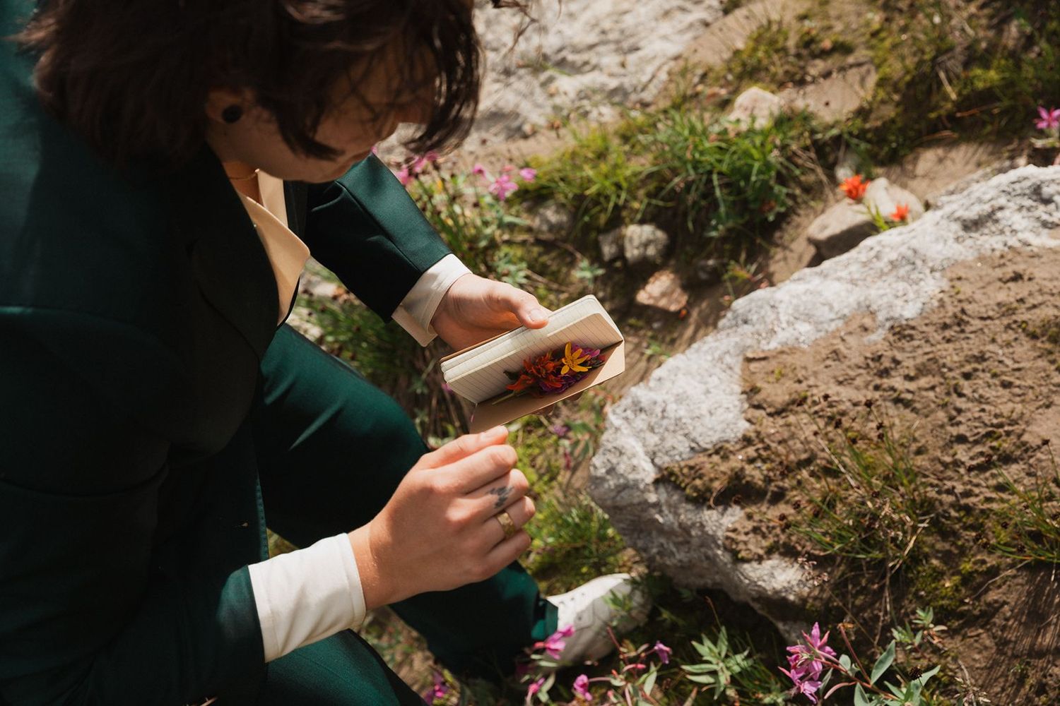 Person in green suit reads vows from a decorated card among wildflowers and rocks.