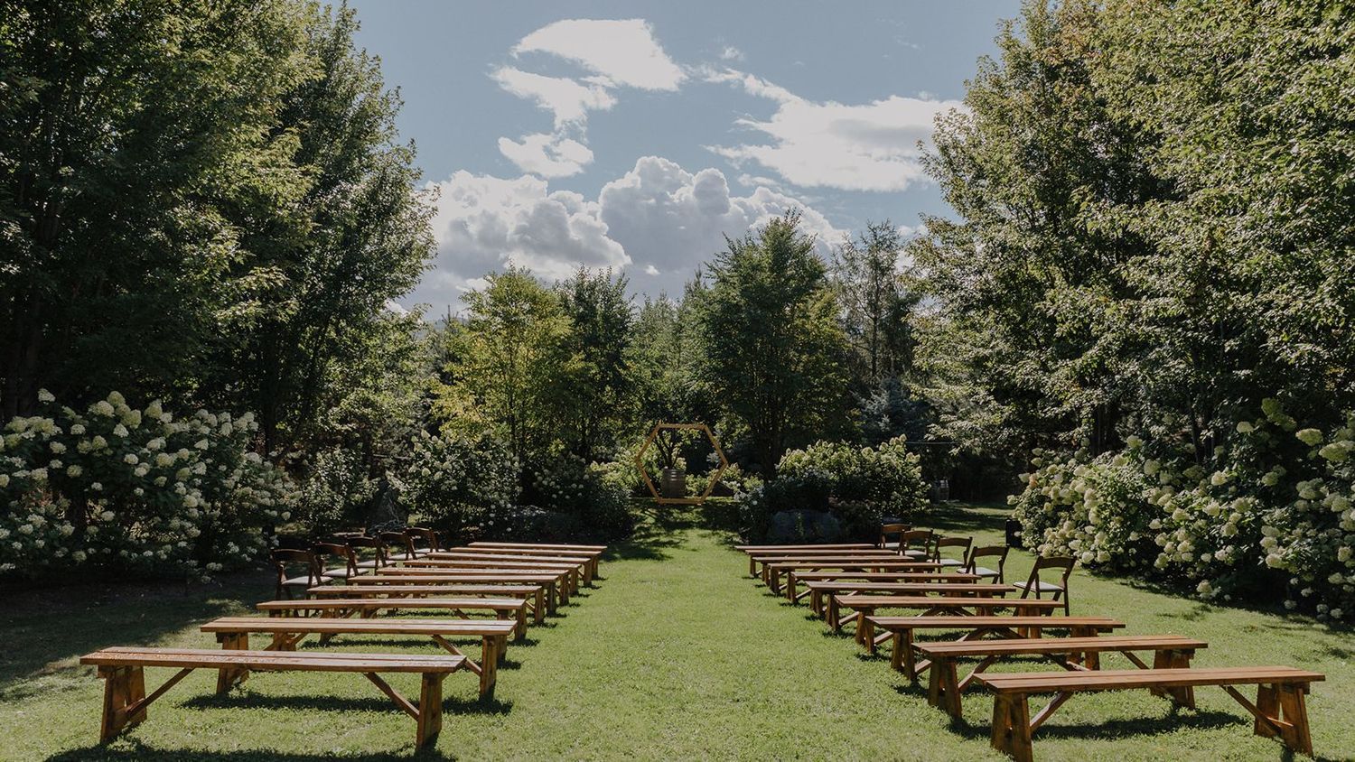 Rows of wooden benches set up for an outdoor wedding ceremony on a lush green lawn surrounded by trees.