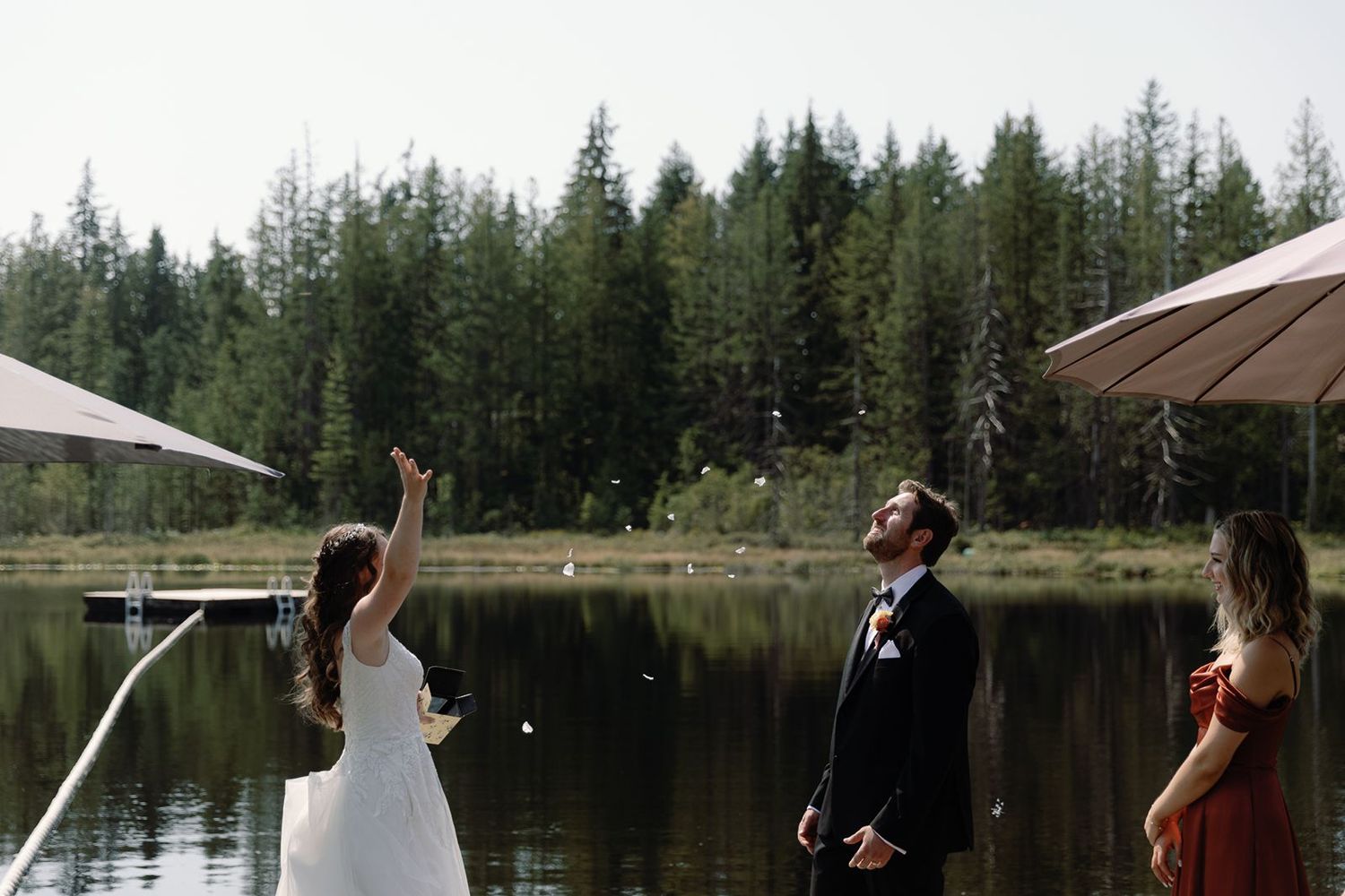 Bride tosses rose petals in the air as groom watches on a scenic lakeside wedding dock.