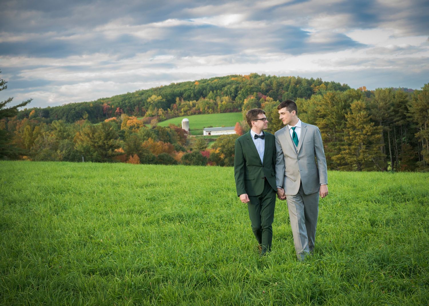 Two grooms in suits walk through a green field with autumn trees and a red barn in the background.