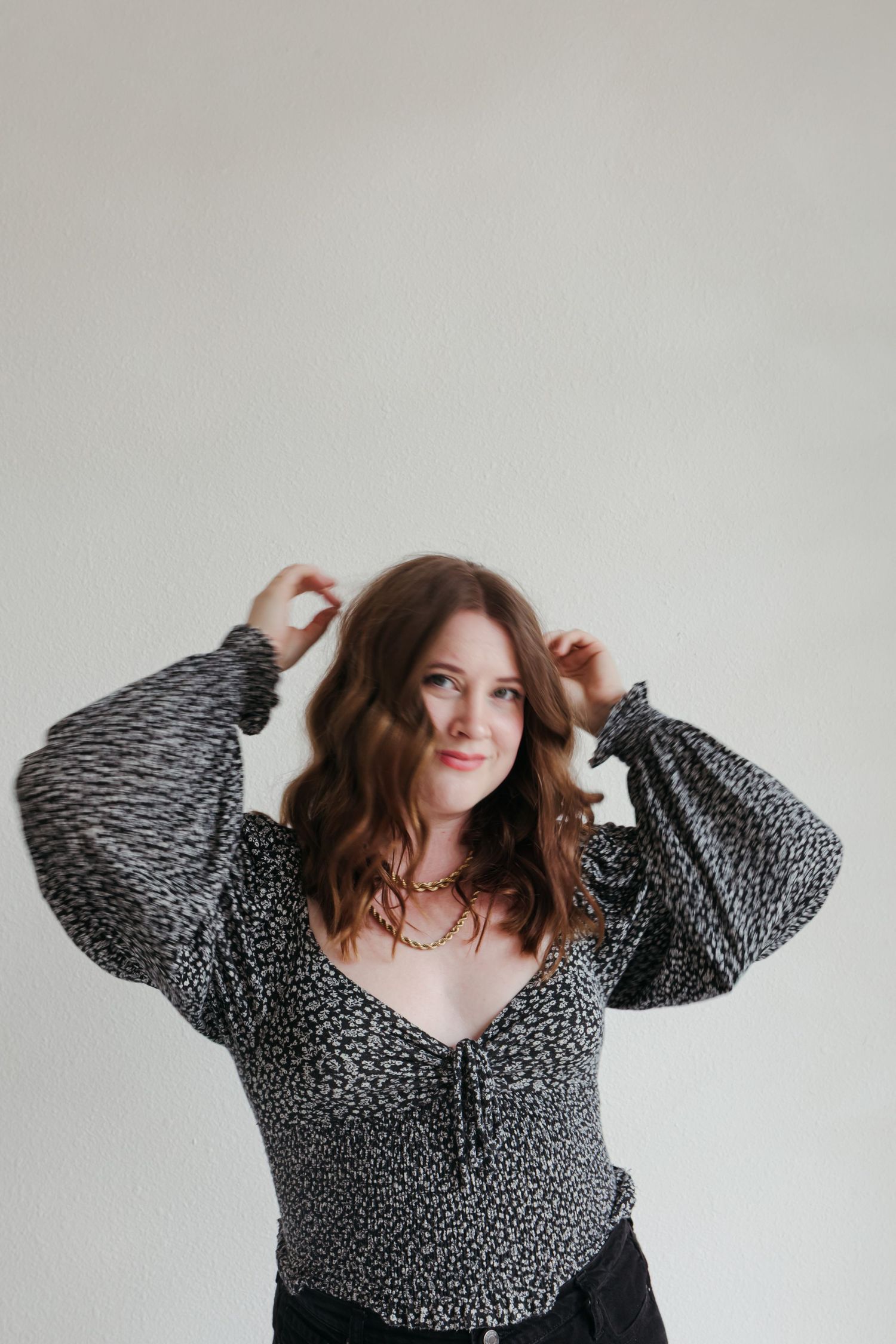 Woman in black and white patterned blouse poses playfully against white wall in fashion photo series.