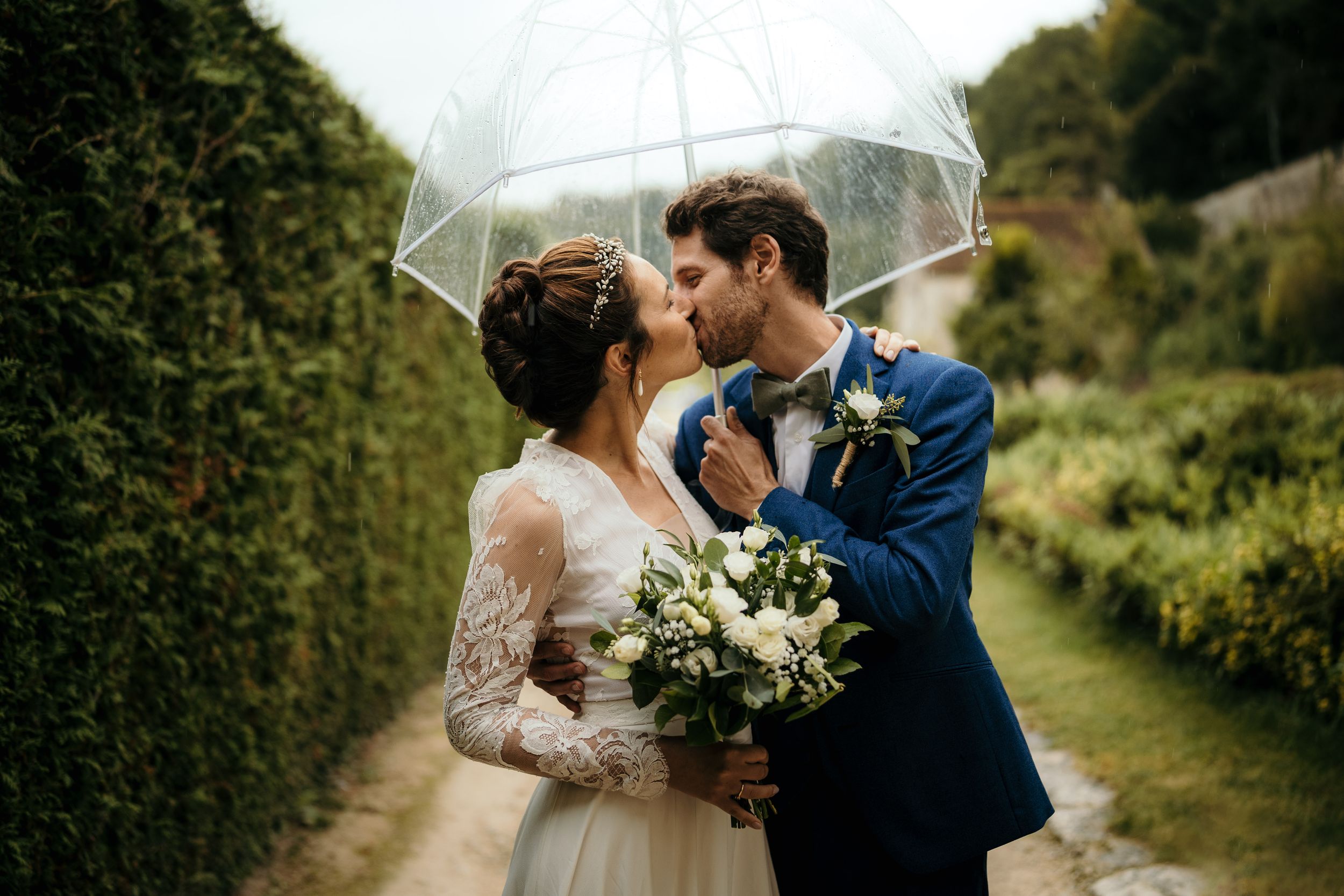 Couple shares romantic moment under clear umbrella on rainy wedding day with white bouquet and navy suit.
