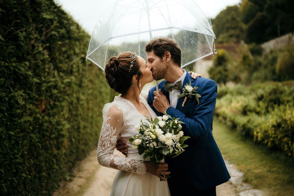 Couple shares romantic moment under clear umbrella on rainy wedding day with white bouquet and navy suit.