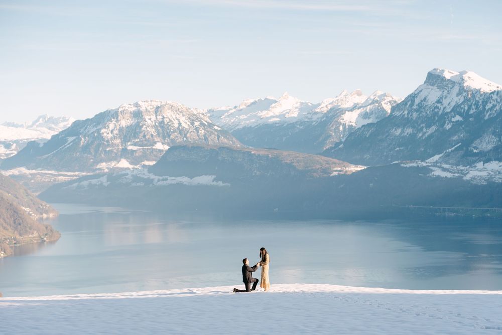 A panoramic view of snow-capped mountains and a lake with two small figures silhouetted in the distance.