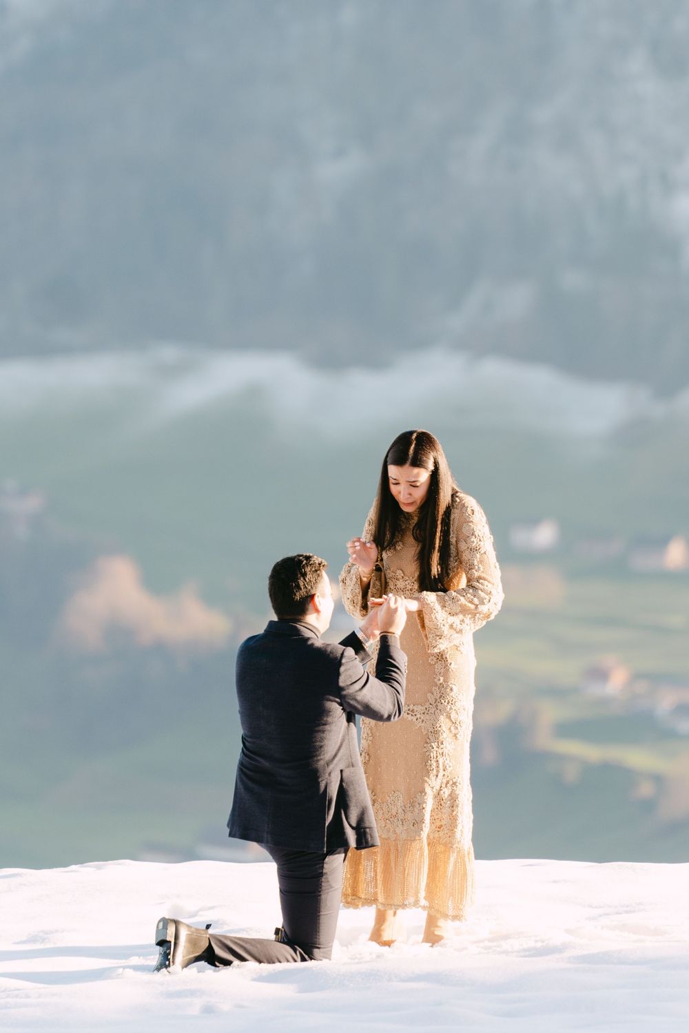 A romantic winter proposal scene on a snowy mountain peak with misty mountains in the background.