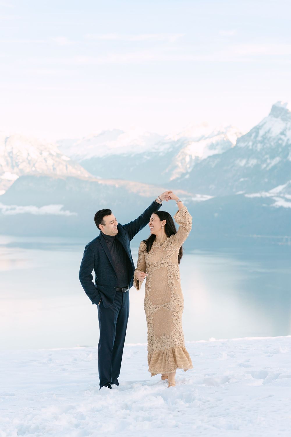 A couple dancing together in formal attire on a snowy mountain overlook with a lake and mountains behind them.