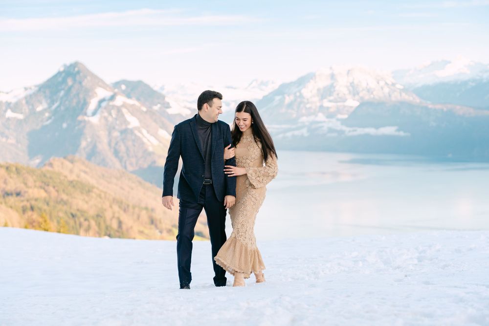 A couple walks together in the snow with majestic snow-capped mountains and a lake in the background.