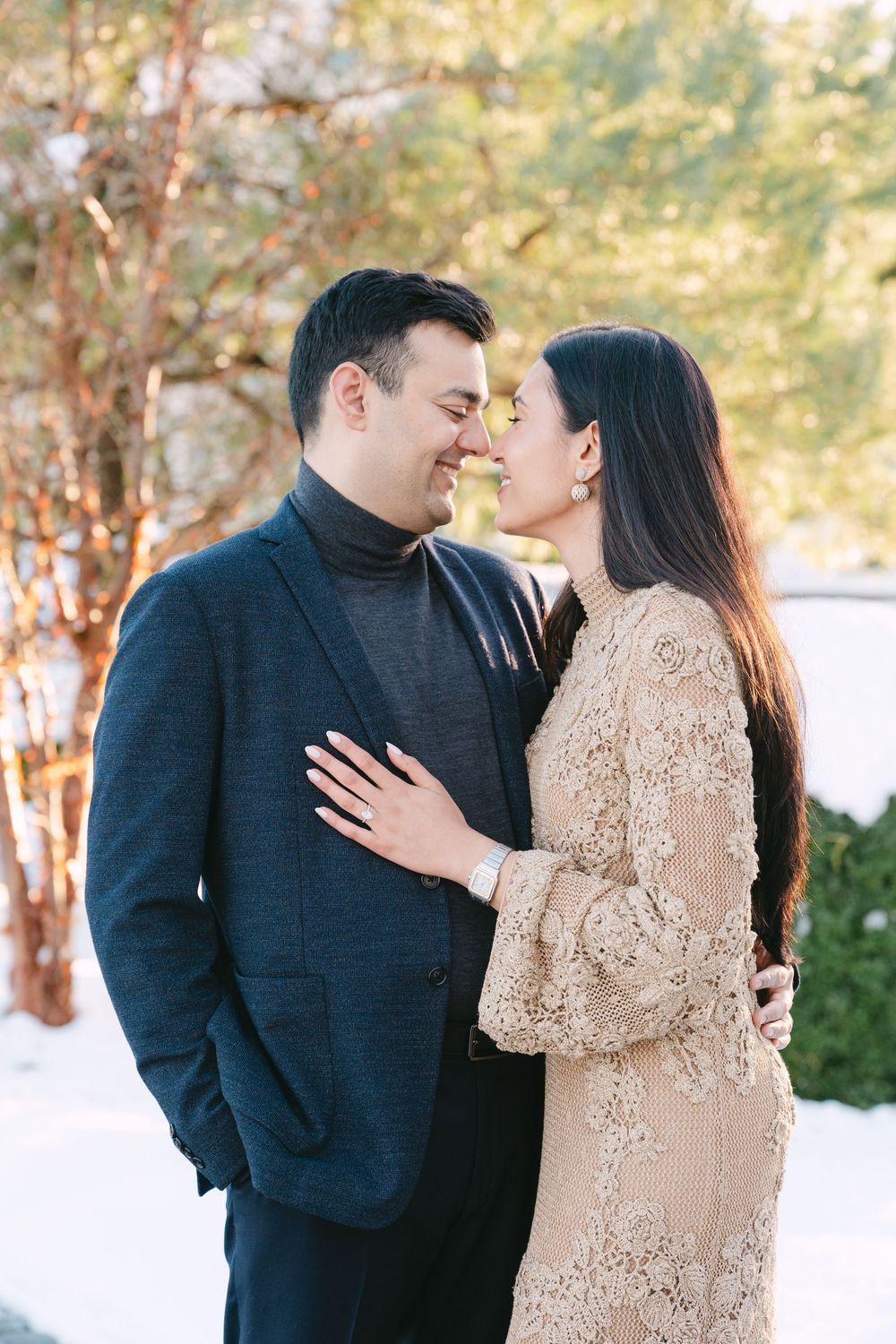 A romantic winter engagement photo showing a couple embracing in front of snowy trees.