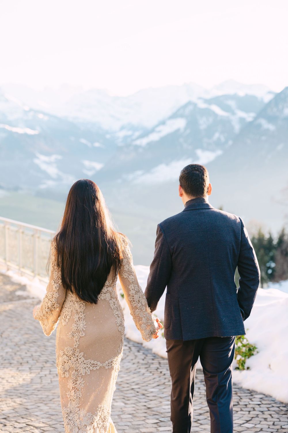 A couple walks hand in hand through snowy mountain scenery during a winter photoshoot.