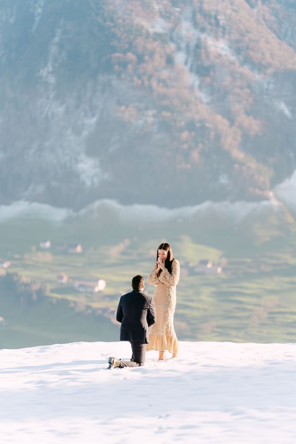 Two figures sitting together on a snowy mountain overlook with dramatic mountain scenery behind them.