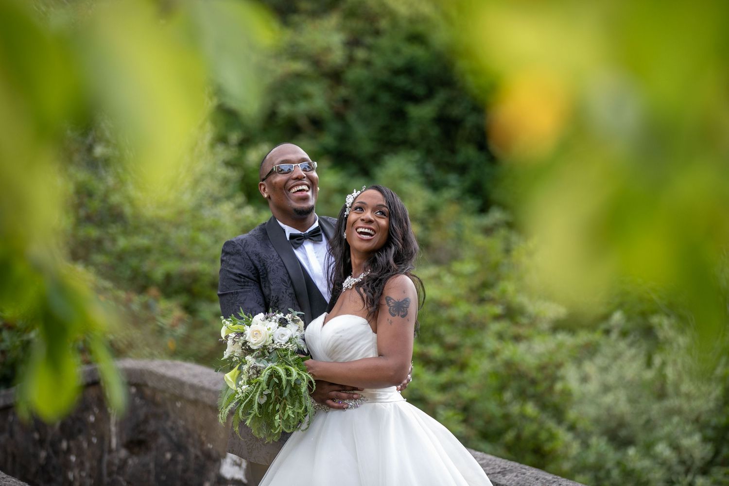 A smiling couple in wedding attire stand on a stone bridge surrounded by lush greenery. The bride holds a bouquet, and both appear joyful