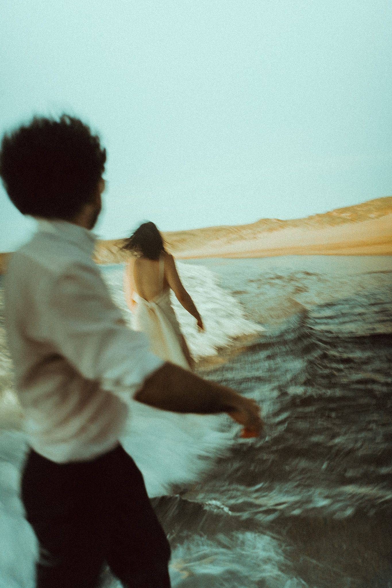 A person in a white shirt stands against a dramatic desert landscape at dusk with rippling sand dunes in the background.