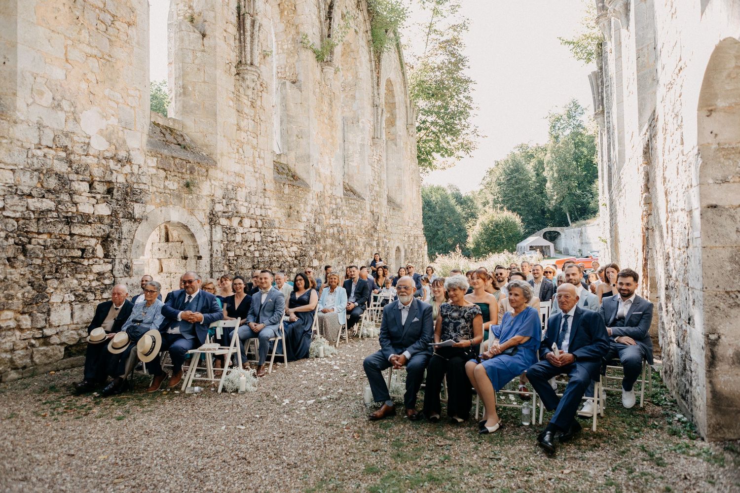 Wedding guests seated in rows within ancient stone abbey ruins during outdoor ceremony on sunny day.