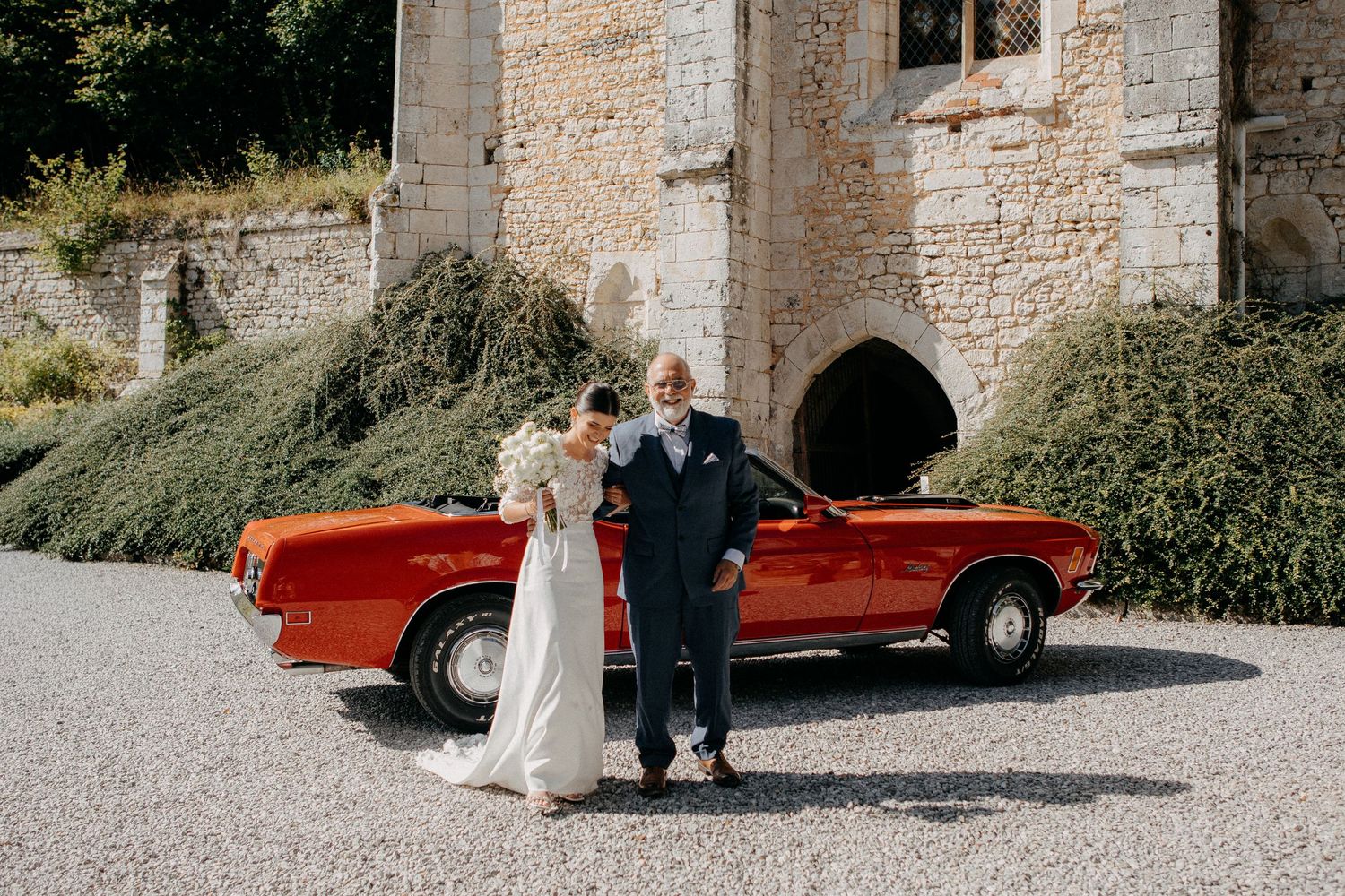 Bride and older gentleman pose together in front of classic red vintage convertible car outside stone castle.