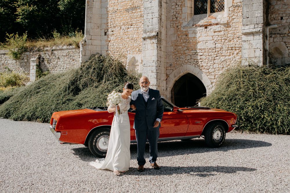 Bride and older gentleman pose together in front of classic red vintage convertible car outside stone castle.