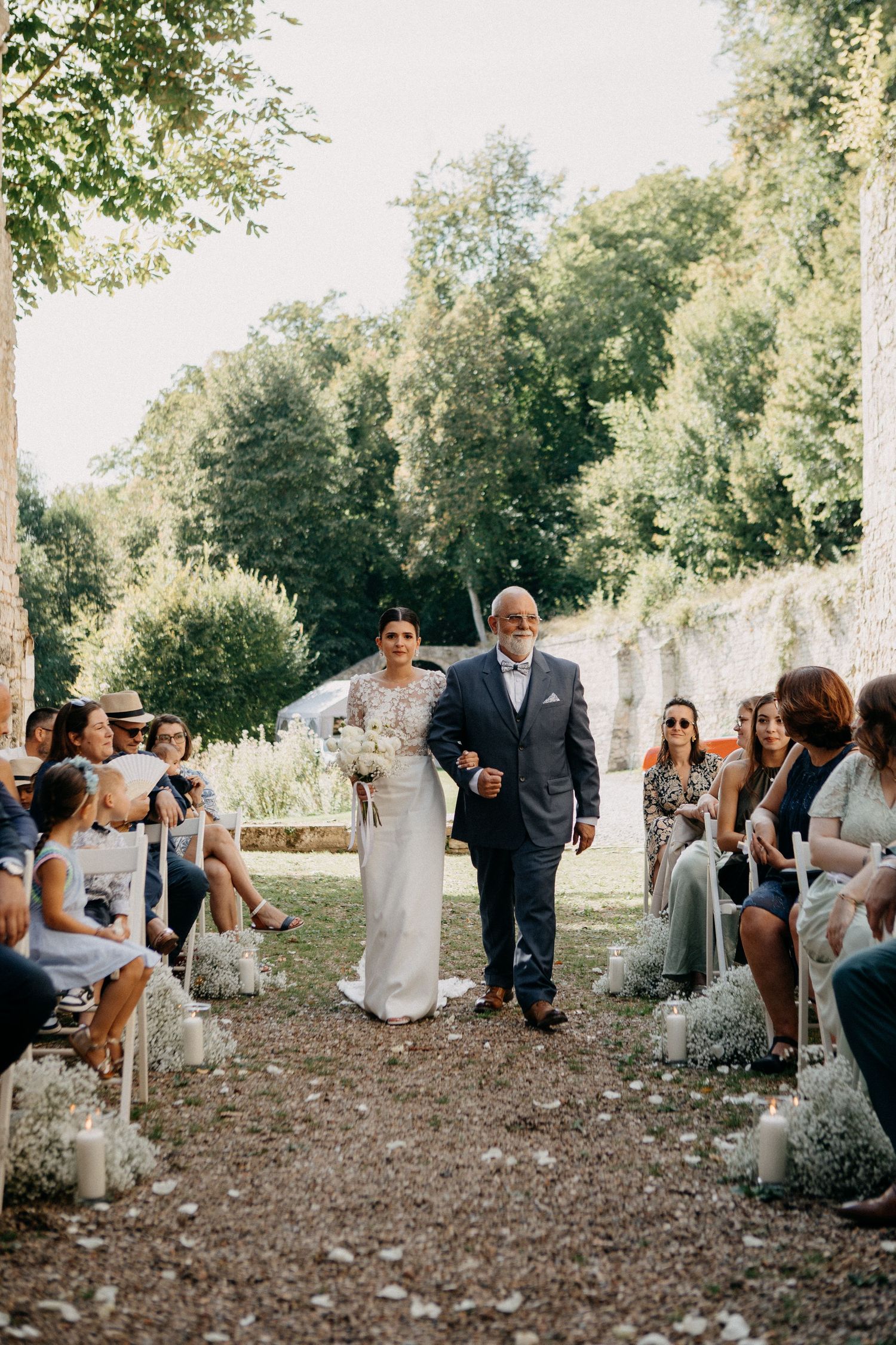Bride in lace gown walks down outdoor aisle lined with candles and gypsophila escorted by father at ceremony.