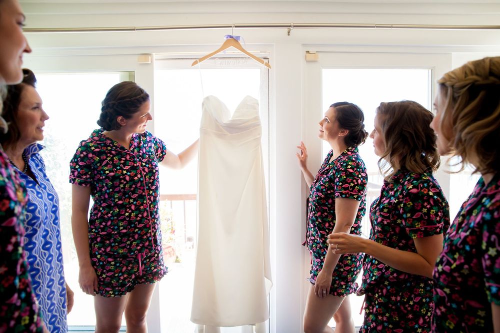 A white wedding dress hangs on display while bridesmaids in floral dresses gather around.