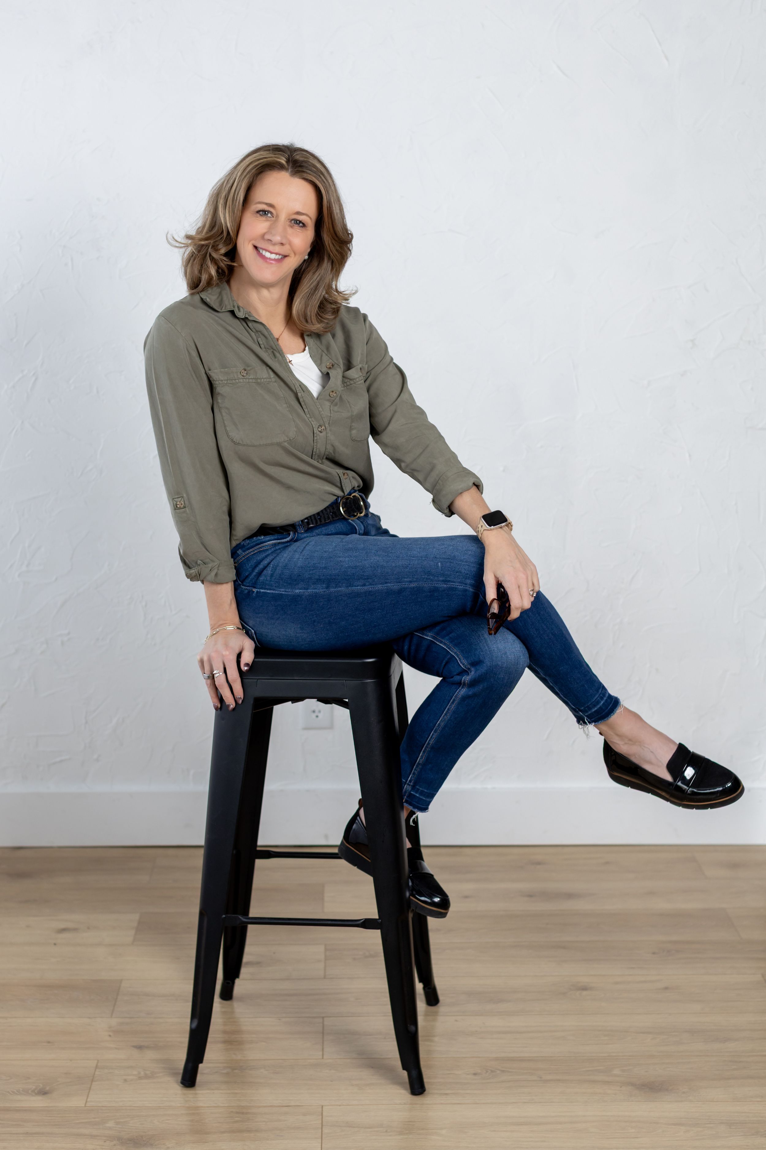 A person in an olive green shirt and blue jeans sits casually on a black bar stool against a white wall.