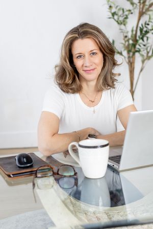 Person in white shirt working at glass desk with laptop and coffee mug in bright minimalist workspace.