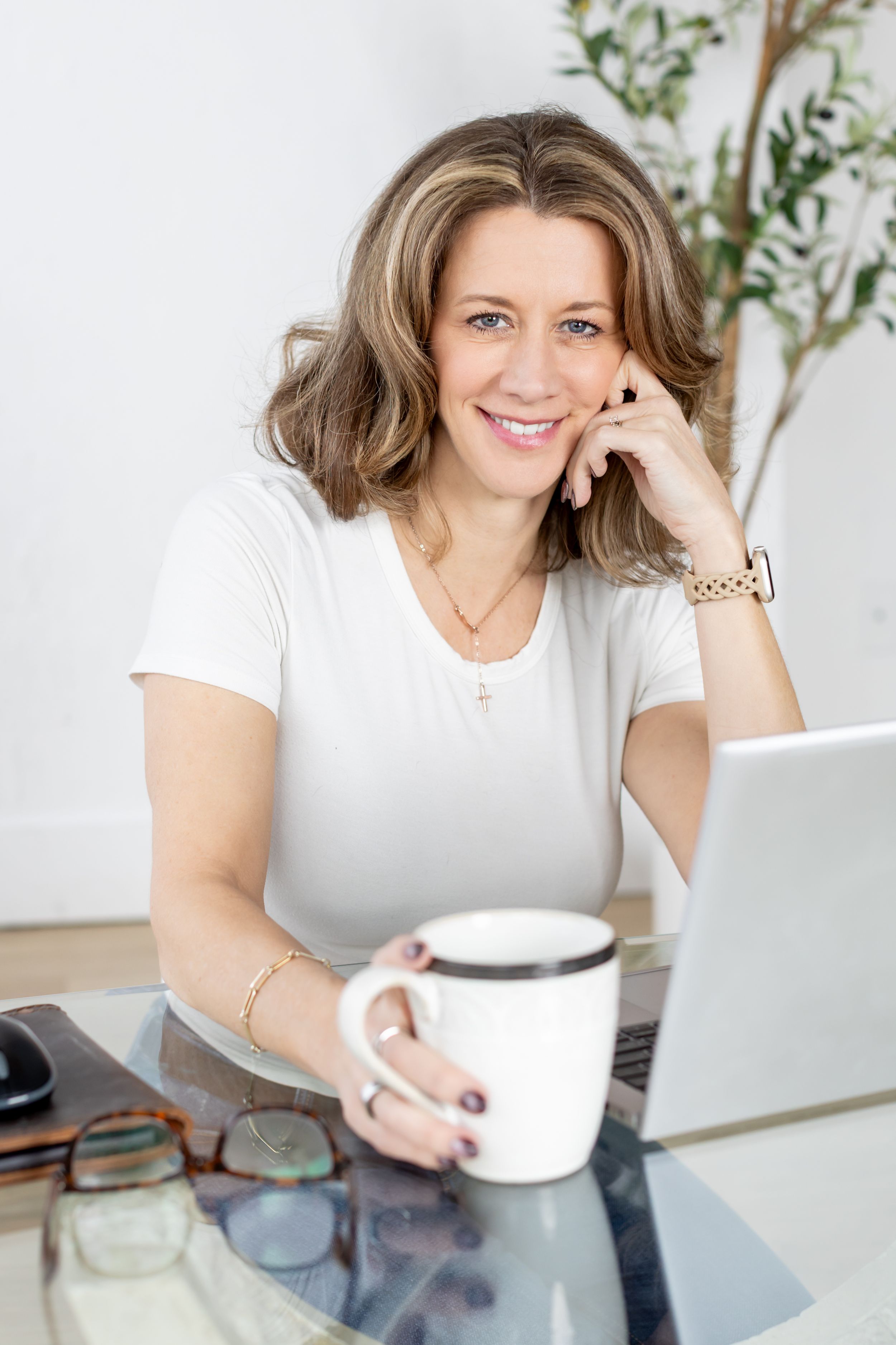 Professional working at desk with coffee cup smiling in bright modern home office with white walls and plants.