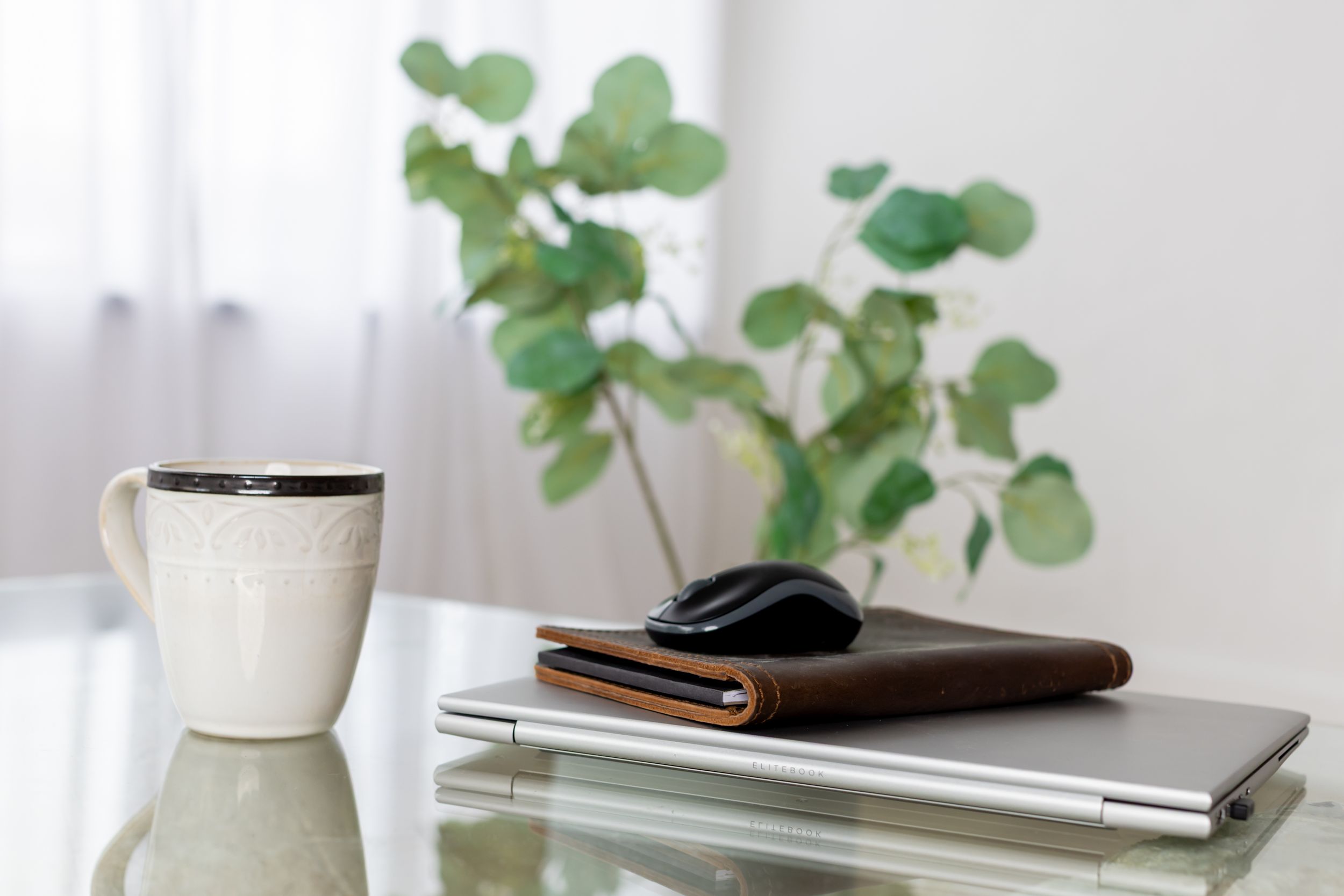 White ceramic vase with eucalyptus leaves sits on a minimalist desk with a laptop and leather notebook.