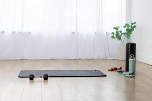 A black yoga mat and fitness equipment laid out on a light wooden floor next to green potted plants.