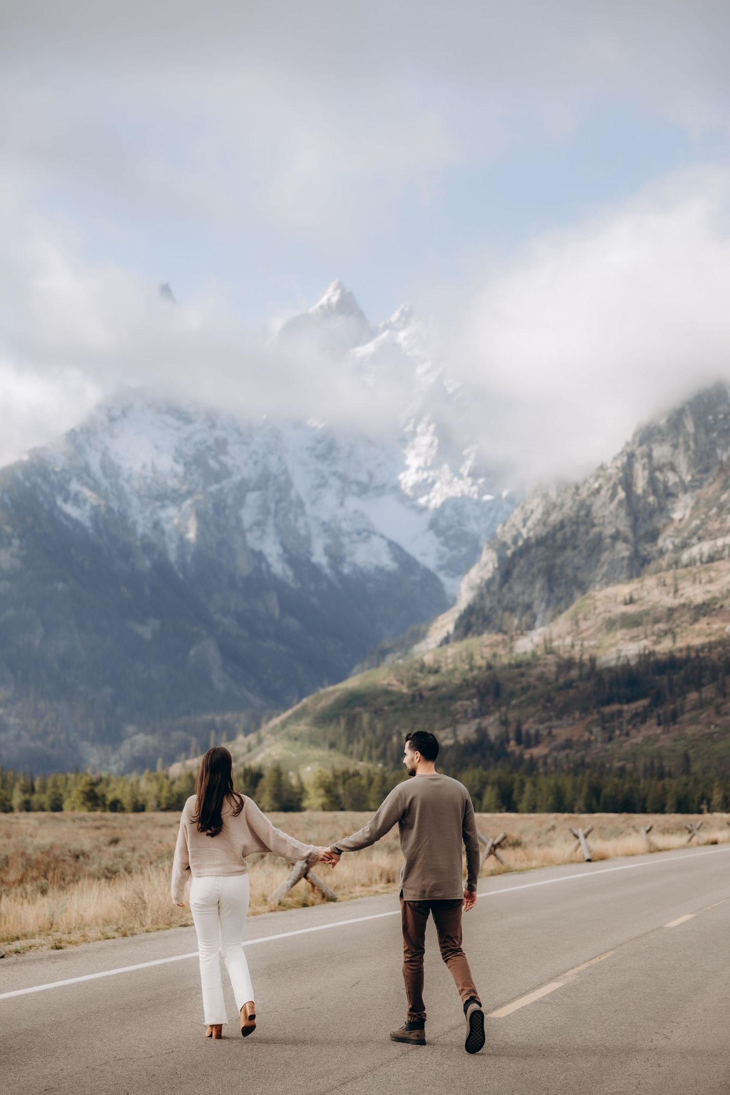 A couple walks along a mountain road with snow-capped peaks in the background on a cloudy day.