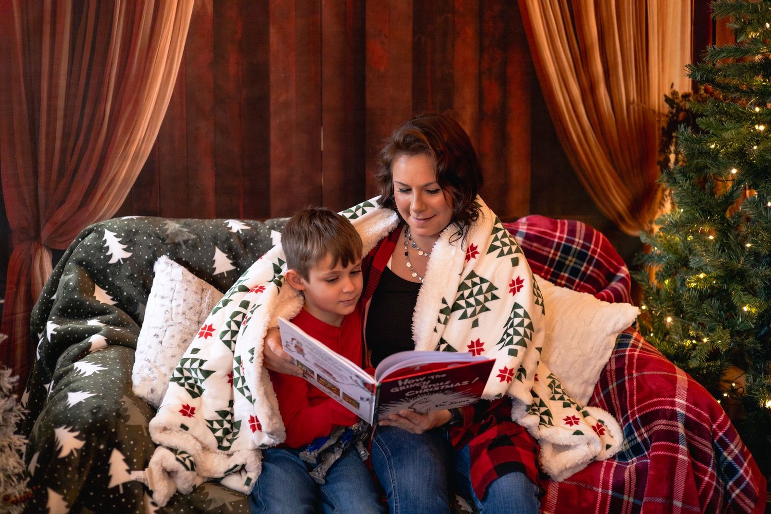 A cozy Christmas scene with people reading a book together on a sofa with plaid blankets and a decorated tree nearby.