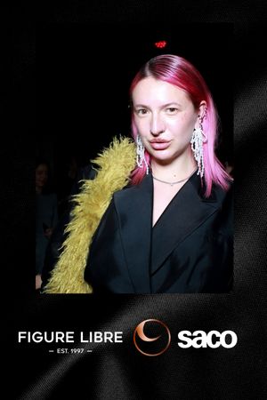 Fashion model wearing black blazer with yellow feather accent and pink hair against dark background at Figure Libre Saco show.