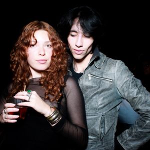 Two people at a nightclub pose together, one with long curly red hair holding a drink and one wearing a gray jacket.