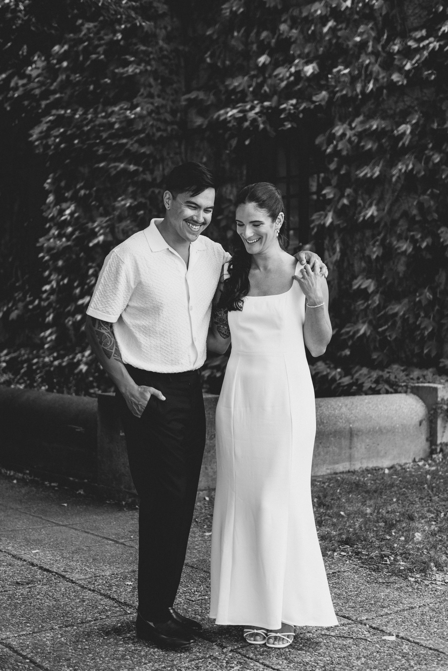 A black and white photo of a couple in formal attire standing together against a leafy backdrop.