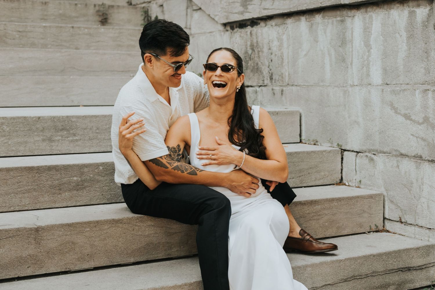 A couple shares joyful moments together while sitting on concrete steps during a sunny photoshoot.