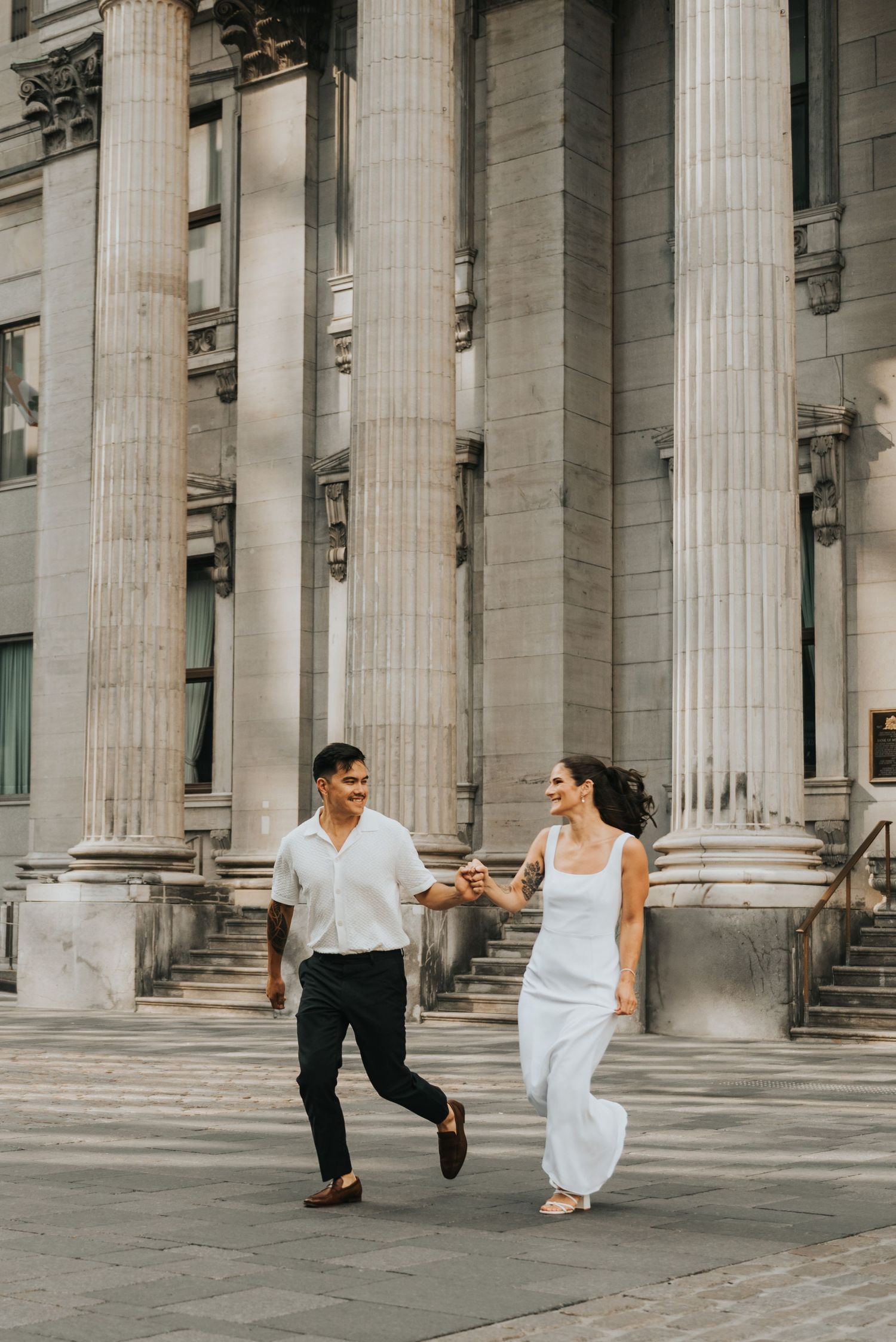 A couple dances playfully between classical stone columns of a grand building with white pillars.
