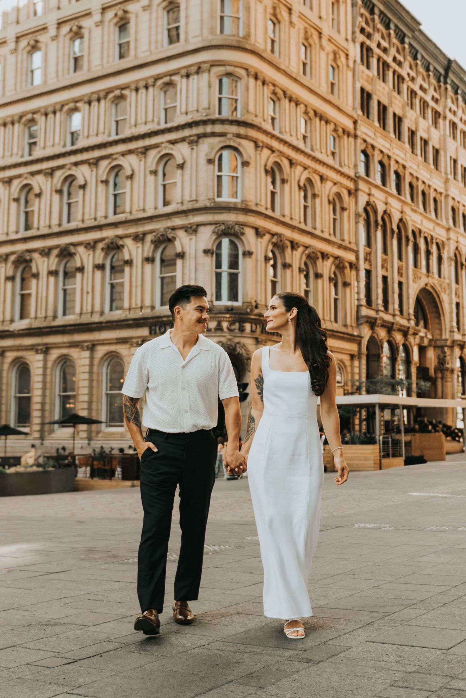 A couple walks hand in hand through historic European city streets with elegant stone architecture in the background.
