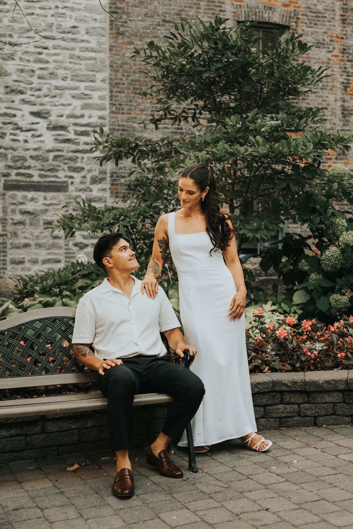 A romantic couple poses together against a stone wall and garden backdrop in elegant white and black attire.