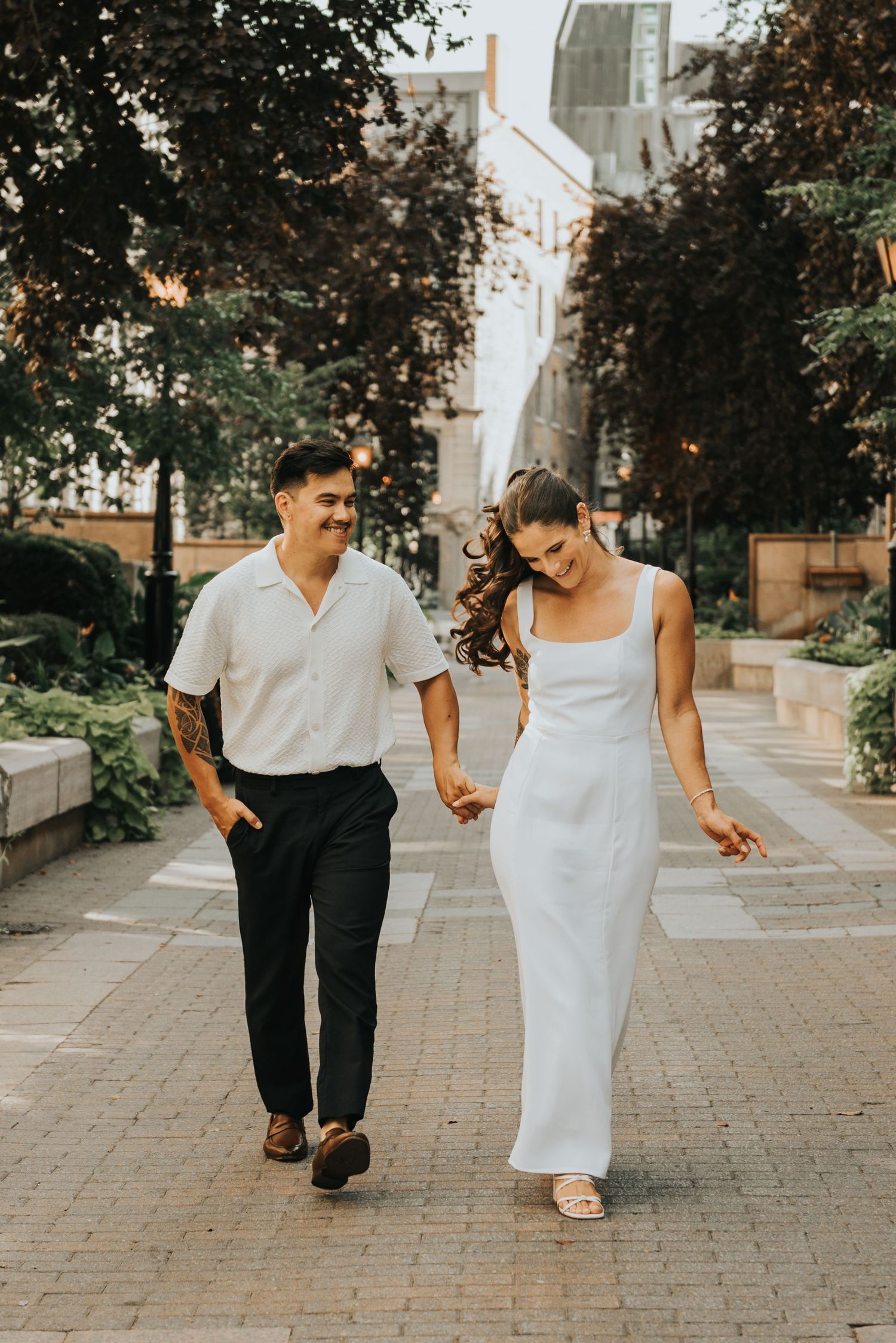 A couple walking hand in hand on a stone pathway surrounded by lush greenery and buildings on a sunny day.