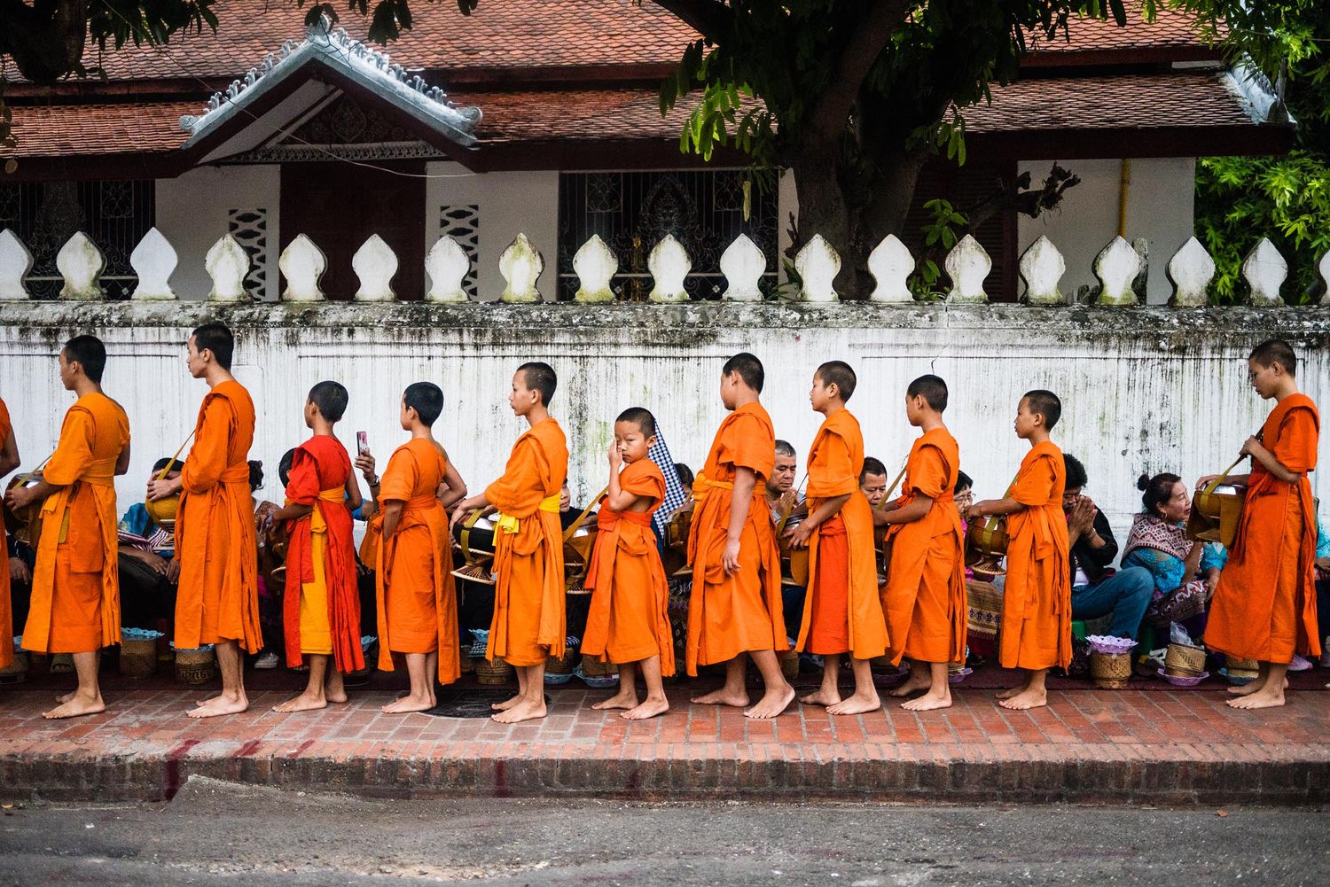 Buddhist monks in bright orange robes stand in a row against a temple wall with white decorative railing.