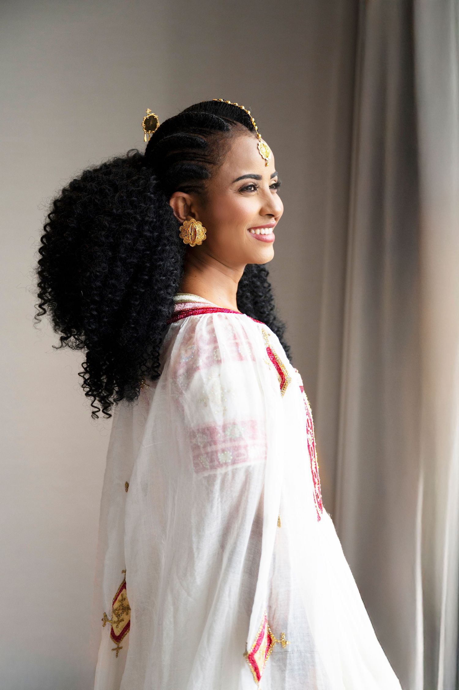 Smiling bride in traditional Ethiopian white habesha kemis dress with gold jewelry and braided curly hair.