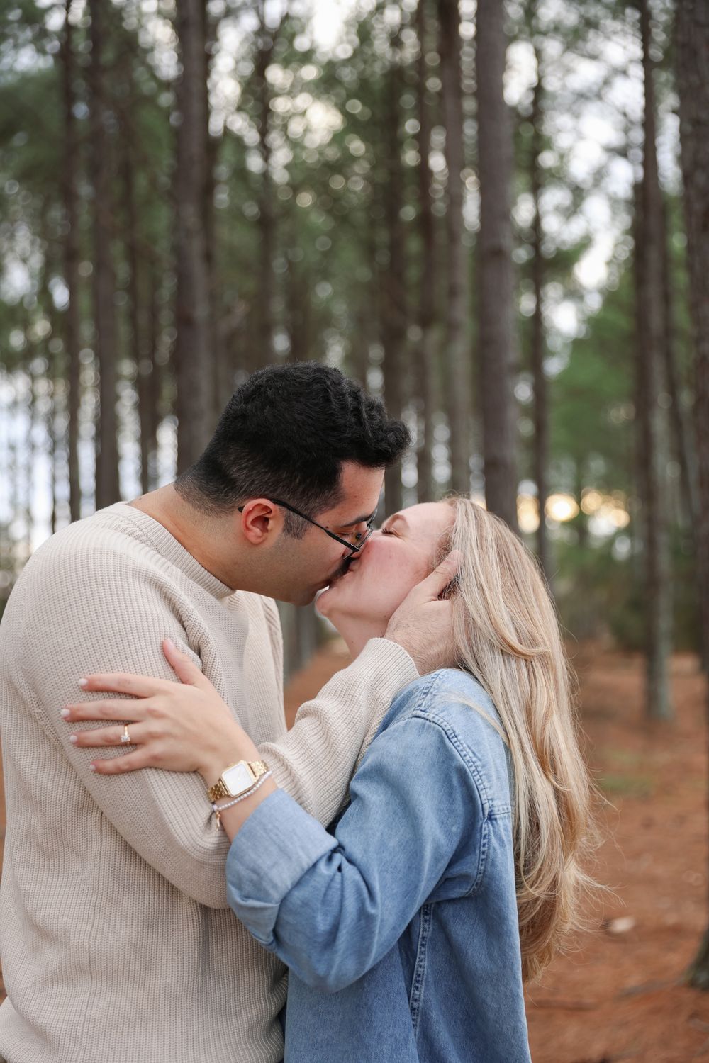 A couple shares a romantic kiss while embracing in a serene forest setting with tall pine trees in the background.