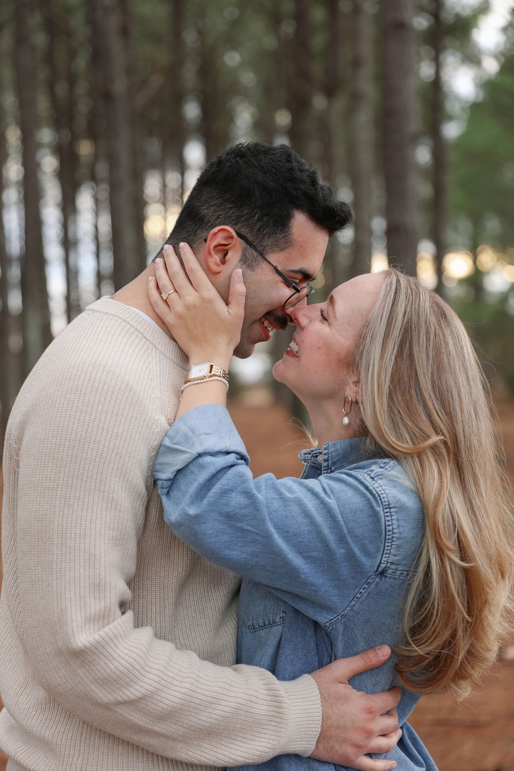 A romantic couple embraces in a pine forest during golden hour wearing casual sweater and denim outfits.