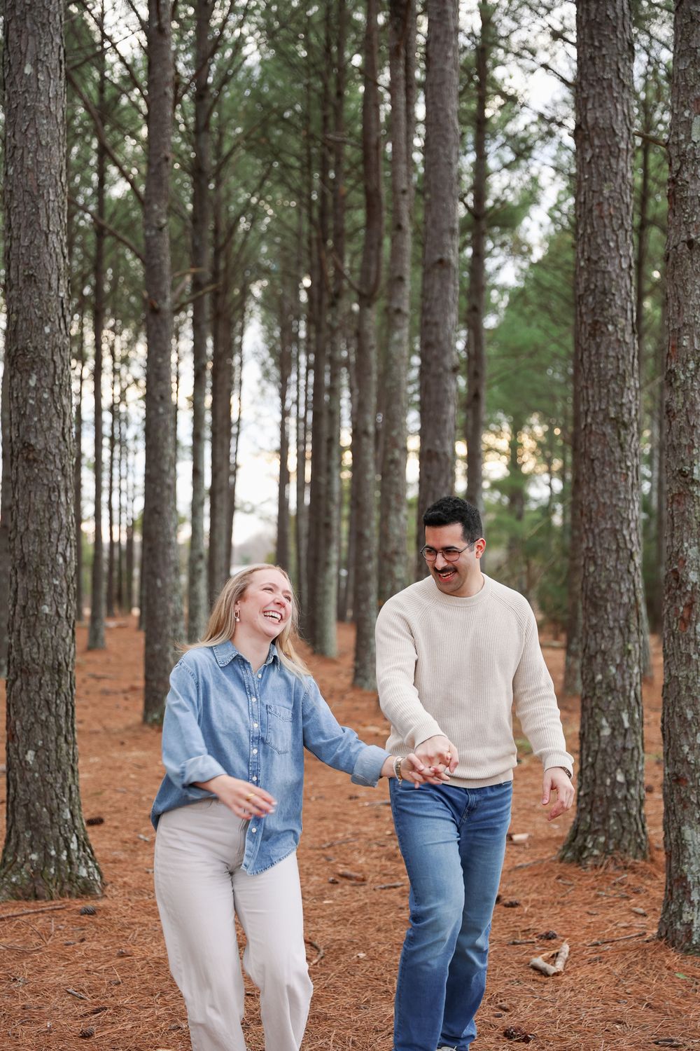 A couple walking and laughing together on a trail through a pine forest with reddish ground cover.