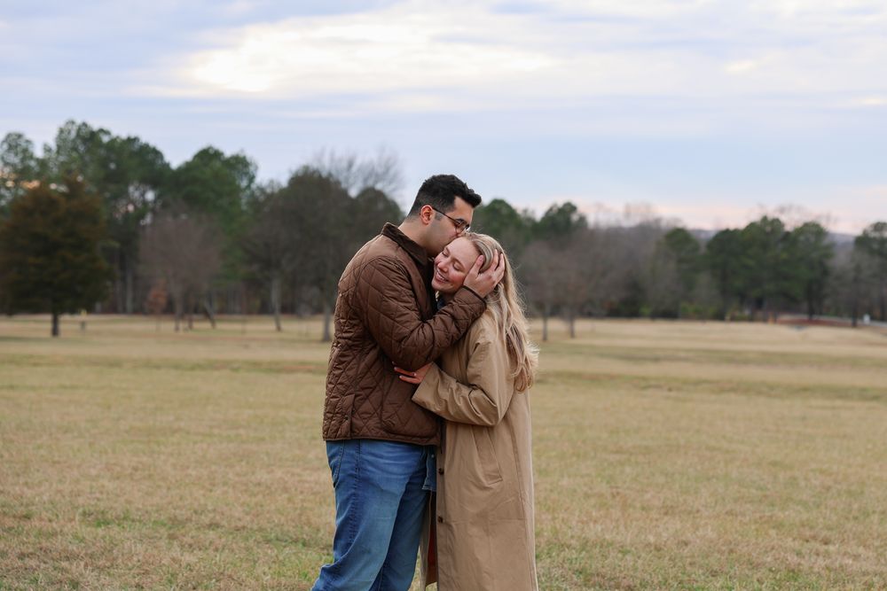A couple shares a romantic moment in an open field with trees in the background during sunset.